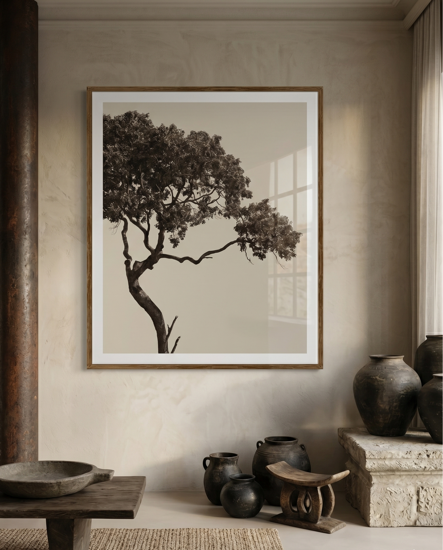 A framed sepia-toned art print of a tree, displayed on a textured beige wall in a styled interior room with rustic pottery and a low wooden table.